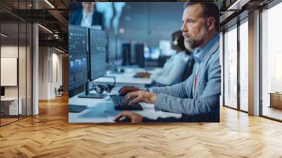 Portrait of Serious Professional Technical Controller Sitting at His Desk with Multiple Computer Displays Before Him. In the Background His Colleagues Working in System Control and Monitoring Center. Wall mural