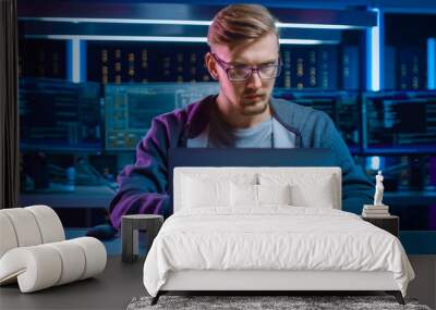 Portrait of Software Developer / Hacker / Gamer Wearing Glasses and Headset Sitting at His Desk and Working / Playing on Laptop. In the Background Dark High Tech Environment with Multiple Displays. Wall mural