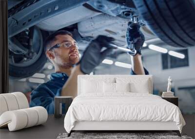 Portrait Shot of a Handsome Mechanic Working on a Vehicle in a Car Service. Professional Repairman is Wearing Gloves and Using a Ratchet Underneath the Car. Modern Clean Workshop.  Wall mural