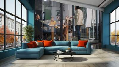 Project Manager Makes a Presentation for a Young Diverse Creative Team in Meeting Room in an Agency. Colleagues Sit Behind Conference Table and Discuss Business Development, User Interface and Design. Wall mural