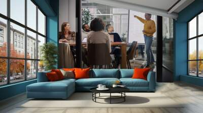 Project Manager Makes a Presentation for a Young Diverse Creative Team in Meeting Room in an Agency. Colleagues Sit Behind Conference Table and Discuss Business Development, User Interface and Design. Wall mural