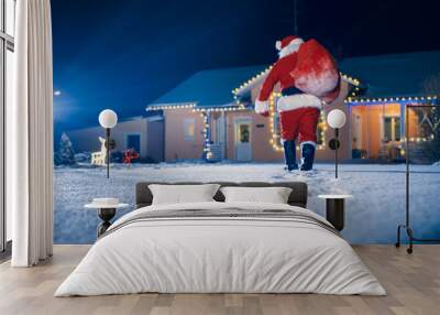 Santa Claus Carrying Red Bag over the Shoulder, Walks into Front Yard of the Idyllic House Decorated with Lights and Garlands. Santa Bringing Gifts and Presents. Wall mural