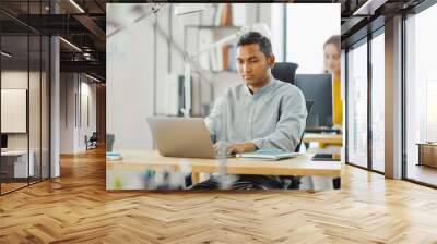 Smart and Handsome Indian Information Technology Specialist Sitting at His Desk works on a Laptop. In the Background Modern Office with Diverse Team of Young Professionals Working. Wall mural