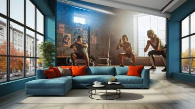 Strong Masculine Man and Two Fit Athletic Women are Doing Squat Exercises. They Workout in a Loft Gym with Motivational Posters on Walls. It's Sunny and Room has Warm Light. Part of Their Team Fitness Wall mural