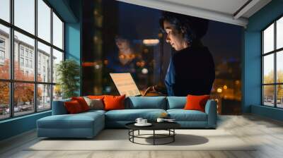 Successful, Stylishly Dressed Businesswoman Holds Laptop While Standing Near the Window of Her Office. Late at Night Professional Woman Doing Important Job. Window Has Big City View with Many Lights. Wall mural