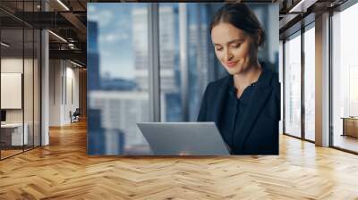 Successful Businesswoman in Stylish Dress Working on Laptop, Standing Next to Window in Big City. Confident Female CEO Analyze Financial Projects. Manager at Work Planning Marketing Campaign. Wall mural