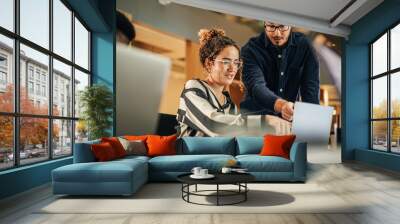 Two Colleagues Working On Laptop in Office. Female Hispanic Help Desk Coordinator Collaborates with White Male Customer Service Agent, They Discuss a Project, Chat, Smile. Teamwork Collaboration Wall mural