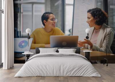 Two Diverse Multiethnic Female Have a Discussion in Meeting Room Behind Glass Walls in an Agency. Creative Director and Project Manager Compare Business Results on Laptop and App Designs in an Office. Wall mural