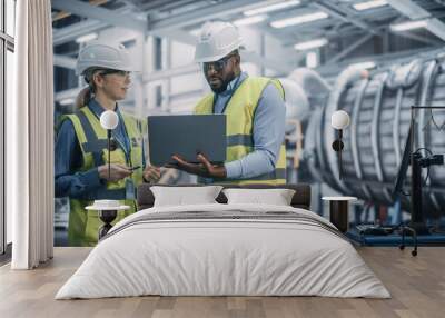 Two Diverse Professional Heavy Industry Engineers Wearing Safety Uniform and Hard Hats Working on Laptop Computer. African American Technician and Female Worker Talking on a Meeting in a Factory. Wall mural