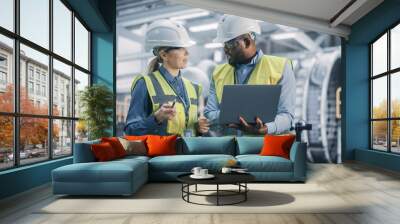 Two Diverse Professional Heavy Industry Engineers Wearing Safety Uniform and Hard Hats Working on Laptop Computer. African American Technician and Female Worker Talking on a Meeting in a Factory. Wall mural