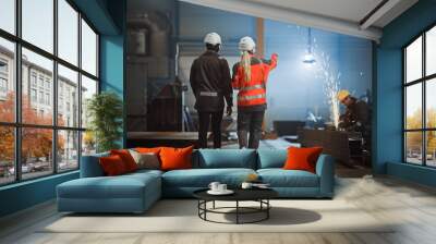 Two Heavy Industry Engineers Walk Away from Camera in Steel Factory and Discuss Work. Industrial Worker Uses Angle Grinder in the Background. African American Specialist Talks to Female Technician. Wall mural