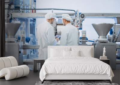 Two Young Food Factory Employees Discuss Work-Related Matters. Male Technician or Quality Manager Uses a Tablet Computer for Work. They Wear White Sanitary Hat and Work Robes. Wall mural