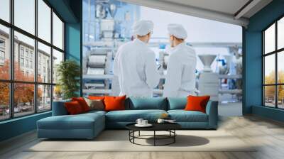 Two Young Food Factory Employees Discuss Work-Related Matters. Male Technician or Quality Manager Uses a Tablet Computer for Work. They Wear White Sanitary Hat and Work Robes. Wall mural