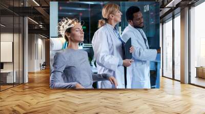 Woman Wearing Brainwave Scanning Headset Sits in a Chair while Two Scientists Supervise and Look at Data. In the Modern Brain Study Laboratory Monitors Show EEG Reading and Brain Model. Wall mural