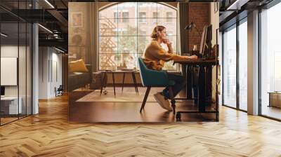 Young Handsome Man Working from Home on Desktop Computer in Sunny Stylish Loft Apartment. Creative Designer Wearing Cozy Yellow Sweater and Headphones. Urban City View from Big Window. Wall mural