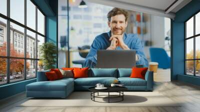 Young Handsome Man Works on a Laptop Computer in Cool Creative Agency in a Loft Office. He has a Take-away Coffee and a Notebook on the Table. He Wears a Jeans Shirt. Wall mural