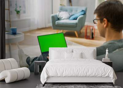 Young Man at Home Works on a Laptop Computer with Green Mock-up Screen. He's Sitting On a Couch in His Cozy Living Room. Over the Shoulder Shot. Wall mural