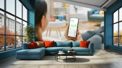 Young Woman at Home Sitting on a Couch and Using Smartphone with Stock Market App to Check Share Prices. Graph is Going Up Making a Great Profit. In the Background Cozy Homely Atmosphere. Wall mural