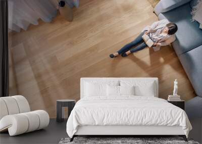 Young Woman is Sitting on a Floor and Reading a Book. Cozy Living Room with Modern Interior, Grey Sofa and Wooden Flooring. Top View Camera Shot. Wall mural