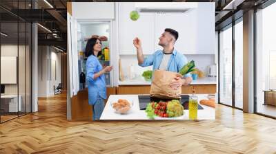 Beautiful young couple unpacking together fresh products from market in the kitchen.The man throws up the cabbage. Wall mural