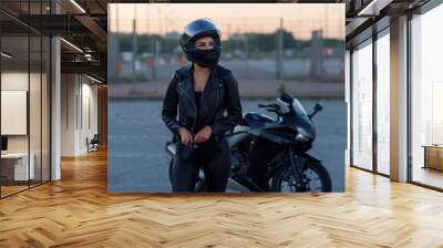 Biker woman in black leather jacket and full face helmet stands near stylish sports motorcycle. Urban parking, sunset in big city. Traveling and active hipster lifestyle. Girls power. Wall mural