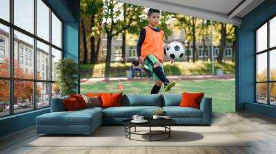 Motivated teen football player stuffs soccer ball on feet with boots. Practicing sport exercises at artificial stadium. Wall mural