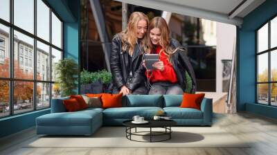 Two stylish girls sitting near shopping center and listening music of watching video on tablet. Wall mural