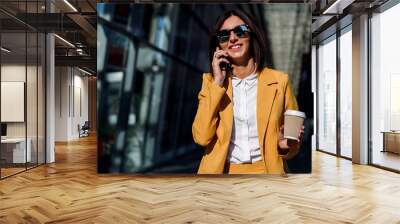 Young smiling business woman in sunglasses and yellow suit stands near office centre with cup of coffee and talks on smart phone. Lunch break among working day. Business lifestyle and success concept. Wall mural