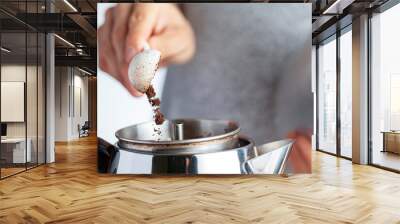 A caucasian woman is taking a teaspoon of ground coffee from a jar using a plastic measuring spoon and putting it into a percolator for brewing homemade morning coffee. Closeup selective focus image. Wall mural