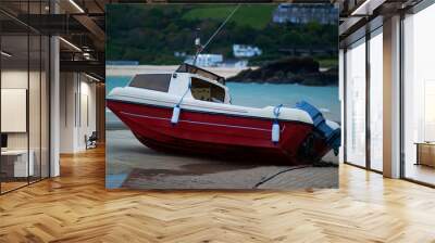 Close up image of a small red and white motor fishing boat that is stranded on shore at low tide. Image was taken at sunset with green hills, sea and buildings in the background. Wall mural