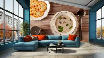 Flat lay image of Creamy mushroom soup decorated with parsley leaves in an artisan porcelain cup on a flat plate on a wooden table served with crackers,  spoon and a salt shaker. Wall mural