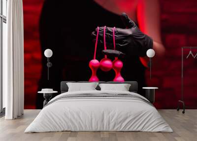 Woman's hand in black latex gloves and a black dress holds pink Kegel balls and Ben Wa balls on a red background. Vaginal balls in a woman's hand. Sex toys Geisha Balls, Products for intimate stores Wall mural