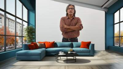 Confident senior man in red casual shirt; blue jeans and eyeglasses posing with arms crossed over white wall background; Business success concept Wall mural