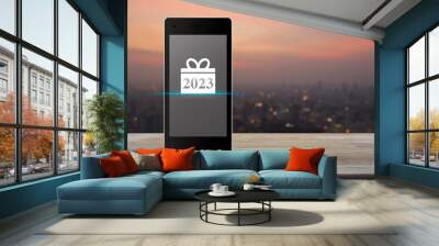 Gift box happy new year 2023 flat icon on modern smart mobile phone screen on wooden table over blur of cityscape on warm light sundown, Business happy new year 2023 shop online concept Wall mural