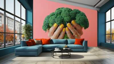 A photo of a woman's hand with yellow nail polish holding broccoli against a pastel pink background Wall mural