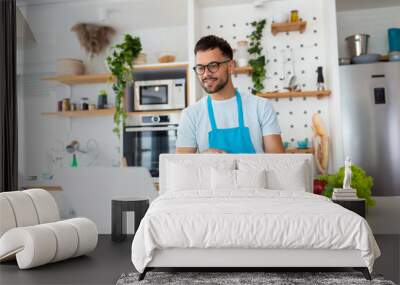 Happy young man cooking healthy dinner at home. He is following a video tutorial on the laptop. Wall mural