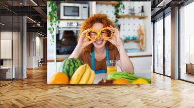 Photo of young woman smiling and holding pepper circles on her eyes while cooking salad with fresh vegetables in kitchen interior at home Wall mural