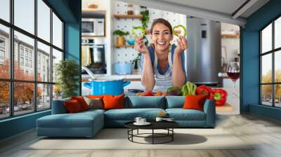 Photo of young woman smiling with green pepper slices while cooking salad with fresh vegetables in kitchen interior at home Wall mural