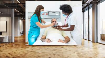 Young doctor and nurse wearing a surgical mask checking on a senior male patient wearing a positive pressure oxygen mask to aid breathing in a hospital bed during the Covid-19 pandemic Wall mural