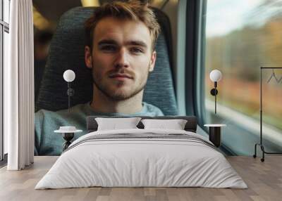 A closeup view of a male traveller with casual attire sitting on a window seat on a moving train, facing the camera, with blurred countryside landscapes passing by.  Wall mural