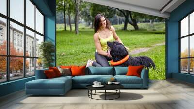 A black briard dog lies on the grass and looks at its owner, who is sitting next to it. Wall mural