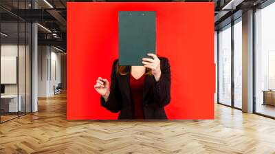 A woman covers her face with clipboard. Human with clipboard in front of his head. Girl manager hides her face. Woman checks list. Woman manager on red background. Businesswoman in black jacket Wall mural