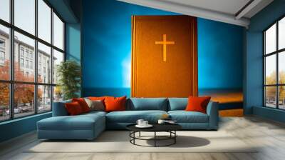 Bible stands vertically on a wooden table. Holy Bible on a blue background. Christian literature. The light falls on the Bible. Reading The Word Of God. Catholicism, Protestantism and Orthodoxy. Wall mural