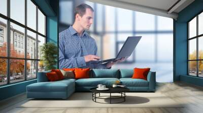 Business man with laptop. Guy is office worker. Man stands with computer in his hands. Manager in business center building. Businessman focused on work. Guy in checkered formal shirt Wall mural