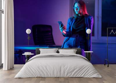 Businesswoman with smartphone in the evening. Woman in the background of the office in dark time. The girl looks at the smartphone screen in neon light. A dark office and a girl with a mobile phone. Wall mural