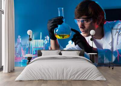 Chemist with a flask in his hands. A man is holding a flask with a yellow chemical liquid. A man in a white coat, goggles, and rubber gloves. Chemical research. Wall mural