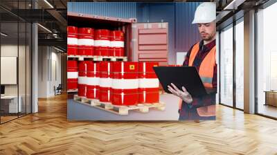 Fuel transportation. Man with laptop in front of shipping container. Barrels of oil on pallets. Red barrels with flammable symbol. Transport sea container with oil. Guy in orange vest. Art focus Wall mural