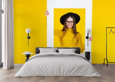 Happy woman holds a white frame in front of her face. Smiling girl. Girl in a yellow dress and a black hat. The lady poses as in the picture. A bright studio shot of a long-haired positive model. Wall mural