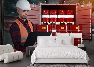 Logistician in warehouse of oil products. Barrels of fuel next to man. Male logistician standing working on laptop. Sea container is filled with barrels of oil. Preparation of oil for export. Wall mural