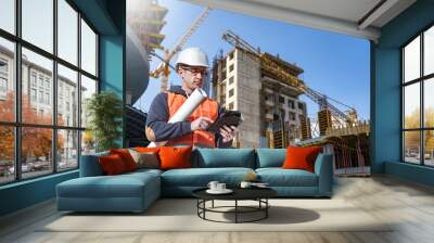 Man builder. Architect at construction site. Builder with tablet computer and papers. Man inspector checks quality of construction. Engineer near multi-story building under construction. Wall mural